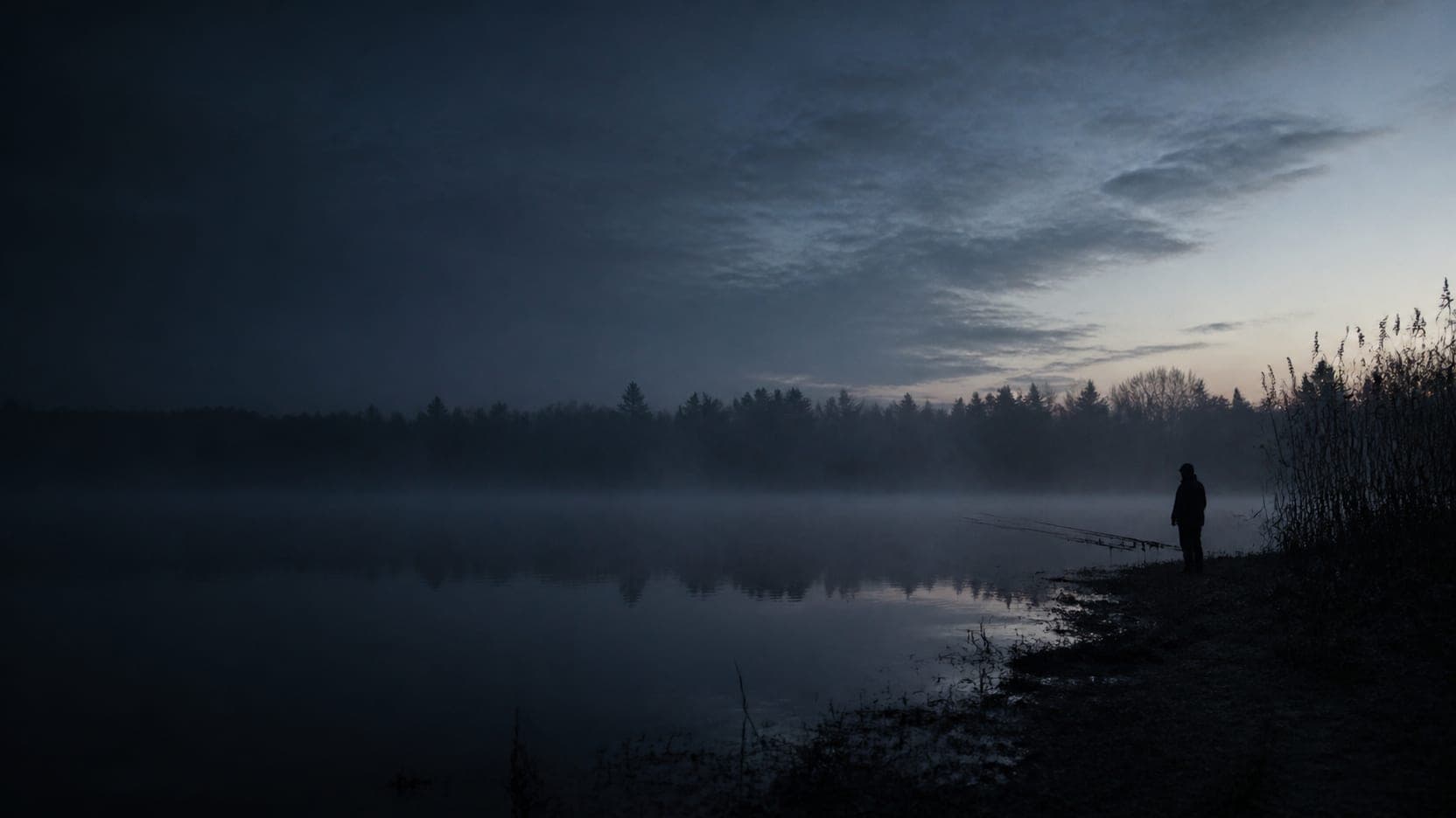 Sylwetka wędkarza na brzegu jeziora o świcie, mgła nad wodą